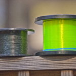 Two spools of fishing line placed on a wooden surface, one with dark green braided line and the other with bright yellow monofilament line, highlighted against a soft blurred background